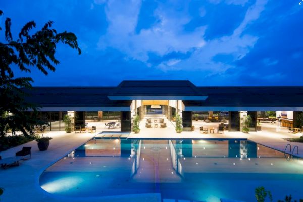 A luxurious outdoor pool in front of a modern, well-lit building during twilight, with clear skies and lush greenery surrounding the scene.