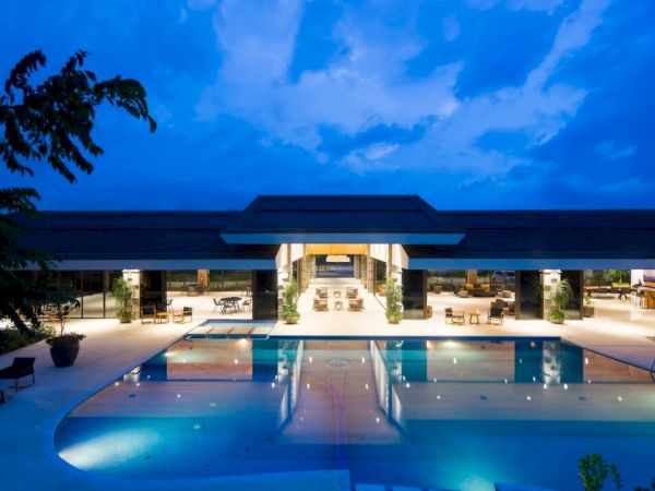 A luxurious outdoor pool in front of a modern, well-lit building during twilight, with clear skies and lush greenery surrounding the scene.