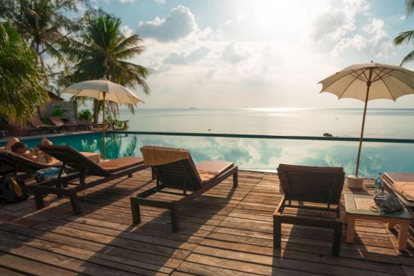 Lounge chairs and umbrellas by a pool with a scenic ocean view, surrounded by palm trees under a partly cloudy sky.