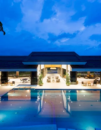 A modern building with a large illuminated swimming pool at night, reflecting the sky and surrounded by seating areas and trees.