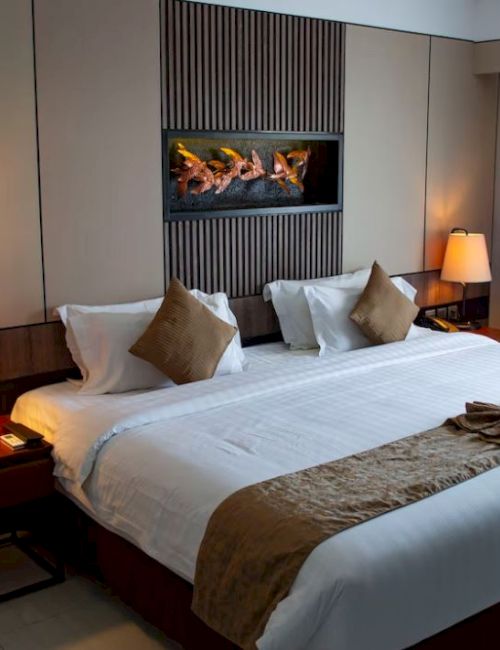 A well-lit hotel room with a large bed, brown pillows, bedside tables with lamps, and a decorative wall feature above the bed.