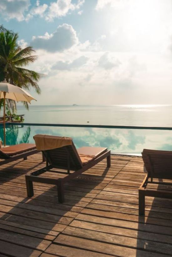 A serene poolside scene with sun loungers, umbrellas, and a view of the ocean under a partly cloudy sky.