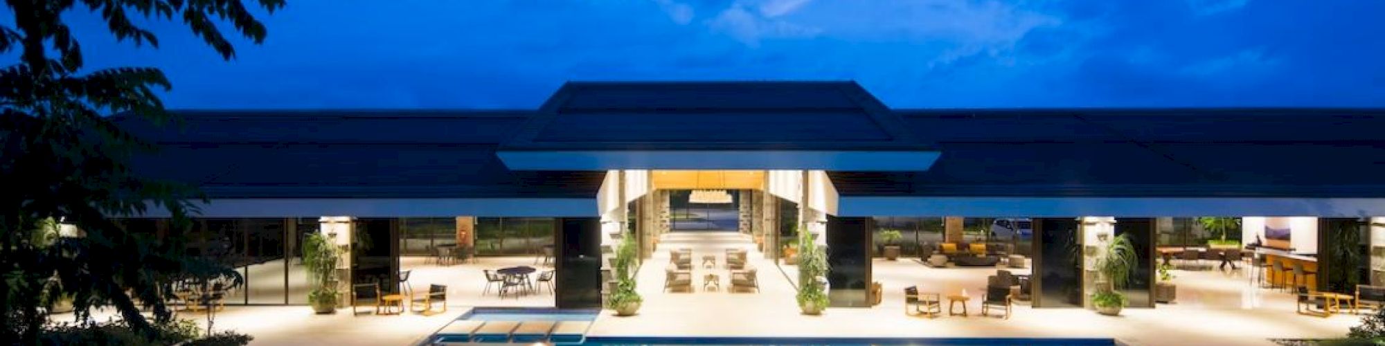 The image shows a modern building with large windows, surrounded by a pool reflecting the lights, under a blue sky with clouds.
