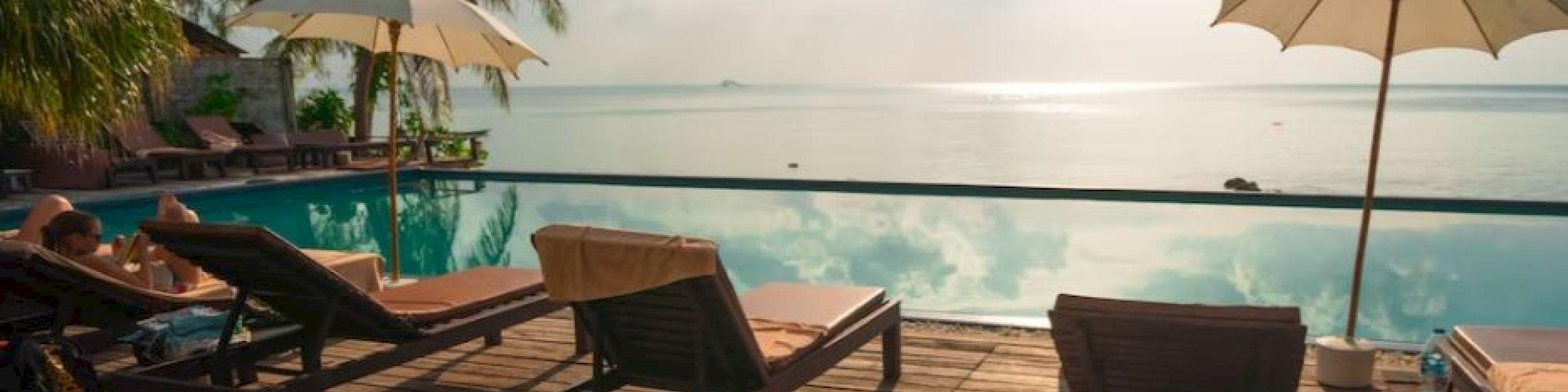 A tranquil poolside scene with lounge chairs, umbrellas, and palm trees overlooking a calm body of water under a partly cloudy sky.