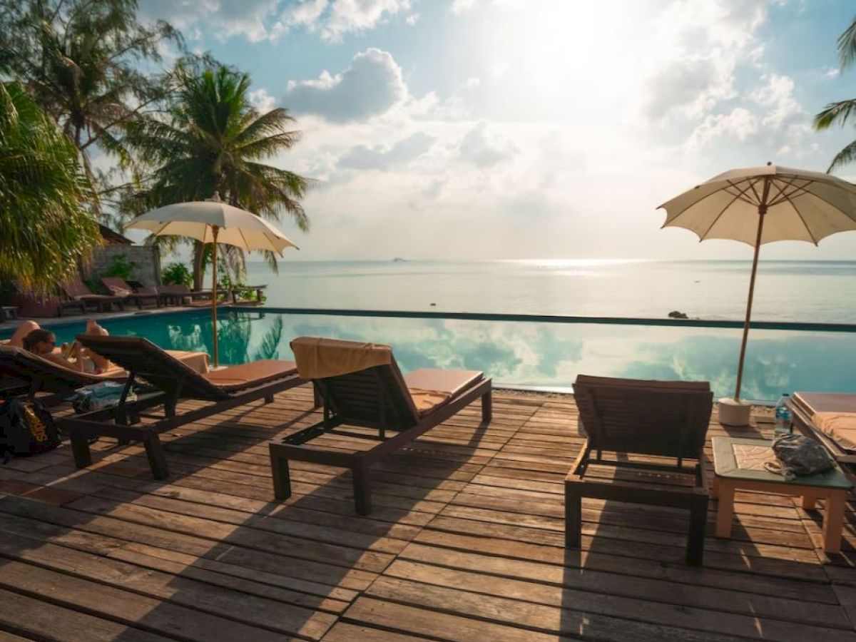Poolside view with lounge chairs and umbrellas, overlooking a calm sea and cloudy sky, surrounded by palm trees and wooden deck.
