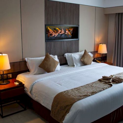 A hotel room with a neatly made king-sized bed, bedside lamps, wall art, and a window with curtains, creating a cozy atmosphere.