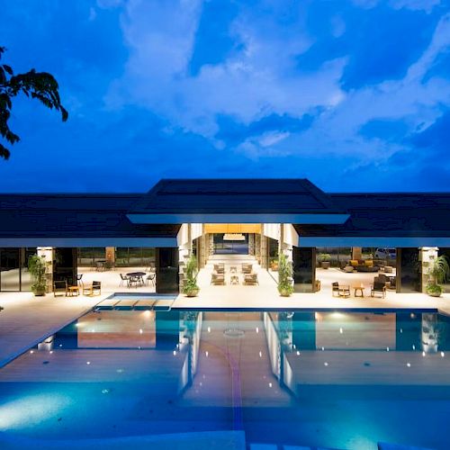 A luxurious building with an illuminated swimming pool at night, featuring open seating areas and surrounded by greenery.