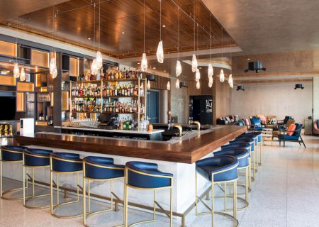 A modern bar with blue stools, a variety of bottles behind the counter, stylish lighting, and a seating area in the background.