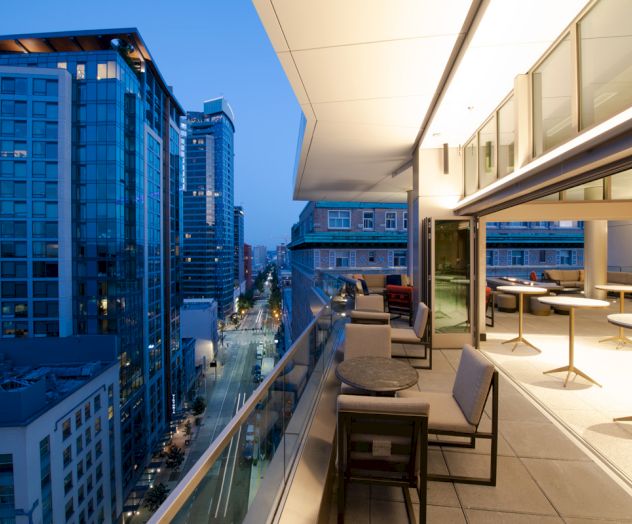 A modern rooftop terrace overlooking a cityscape during dusk, featuring tables, chairs, and a glass railing with lit buildings in the background.