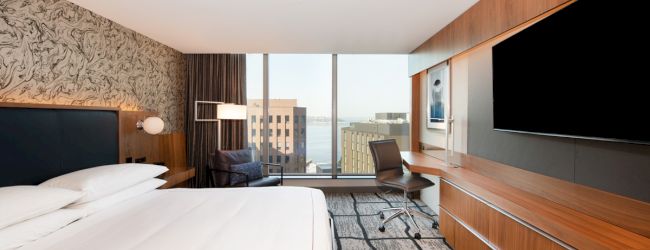A modern hotel room with a large bed, a flat-screen TV, desk, chair, and a window overlooking a cityscape and water.