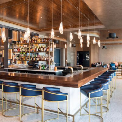 A modern bar with blue chairs, hanging lights, and a well-stocked liquor shelf, surrounded by a spacious seating area.