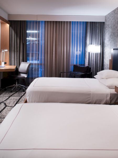 A modern hotel room with two beds, a flat-screen TV, desk, and patterned carpet. The walls have a decorative design, and a window is in the background.