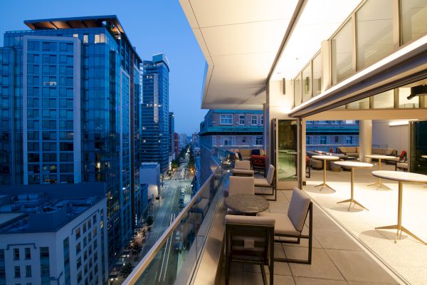 A modern balcony overlooking a cityscape, with tables and chairs, and buildings lit up at dusk under a clear sky.