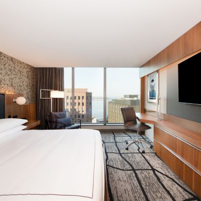A modern hotel room with a large bed, desk, chair, and TV. A window offers a cityscape view. Stylish decor and neutral tones dominate.