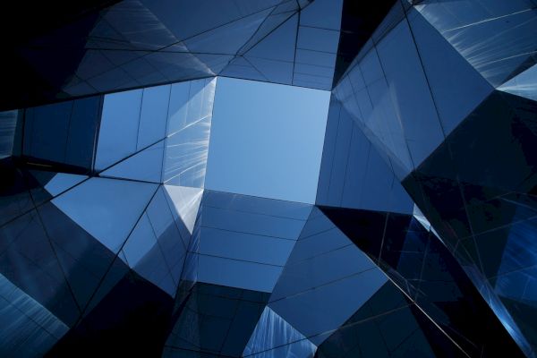 Looking up through abstract, reflective glass structures with a clear blue sky framed in the center, creating a geometric pattern.