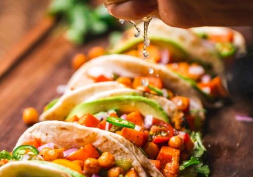 A hand squeezes lime over a row of tacos filled with chickpeas, avocado, and vegetables, garnished with cilantro, on a wooden surface.