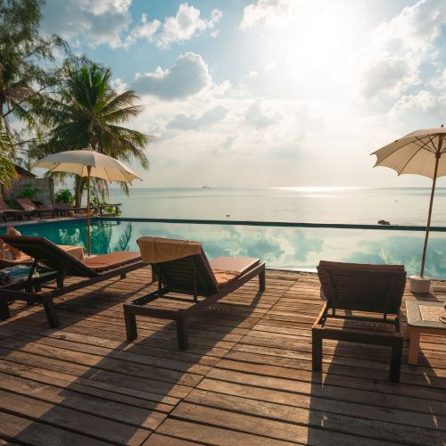 The image shows a tranquil poolside scene with lounge chairs, umbrellas, palm trees, and a view of the ocean under a bright sky.