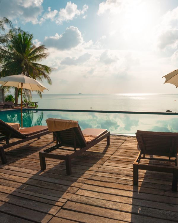 The image shows a tranquil poolside scene with lounge chairs, umbrellas, palm trees, and a view of the ocean under a bright sky.