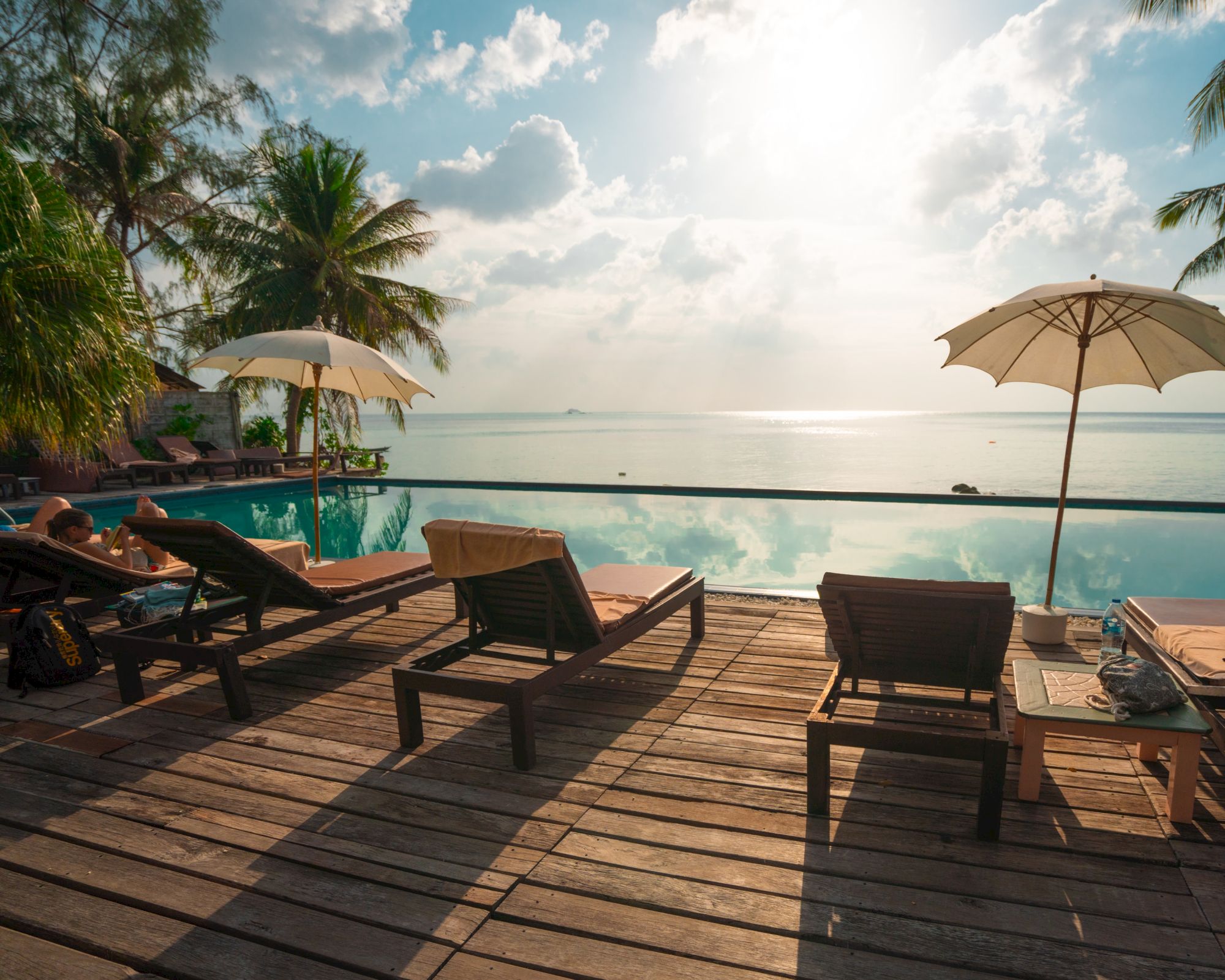 A serene beach scene with lounge chairs, umbrellas, and palm trees by a calm sea under a partly cloudy sky.