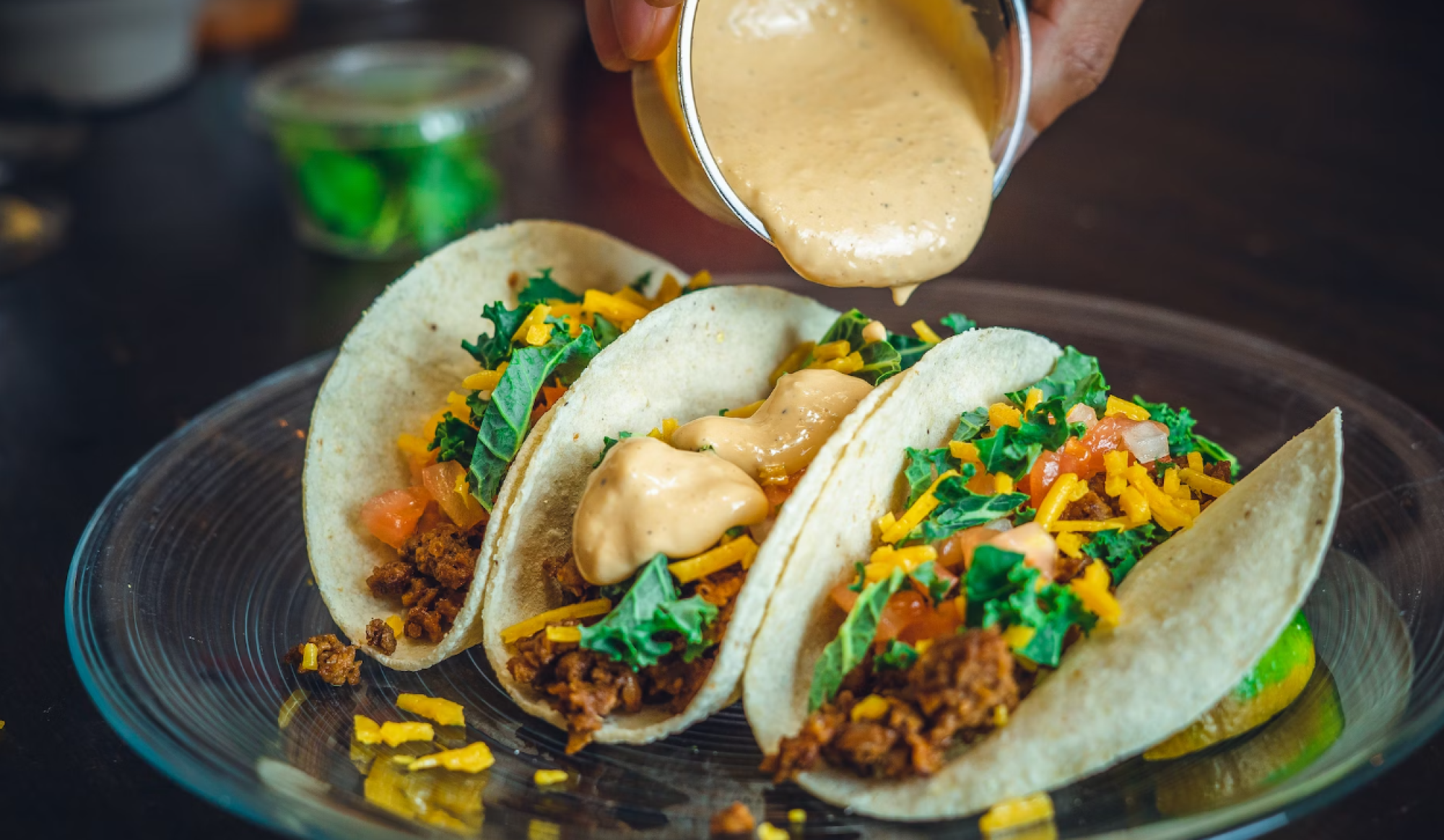 Three tacos filled with meat, cheese, lettuce, and tomatoes are being topped with a creamy sauce.
