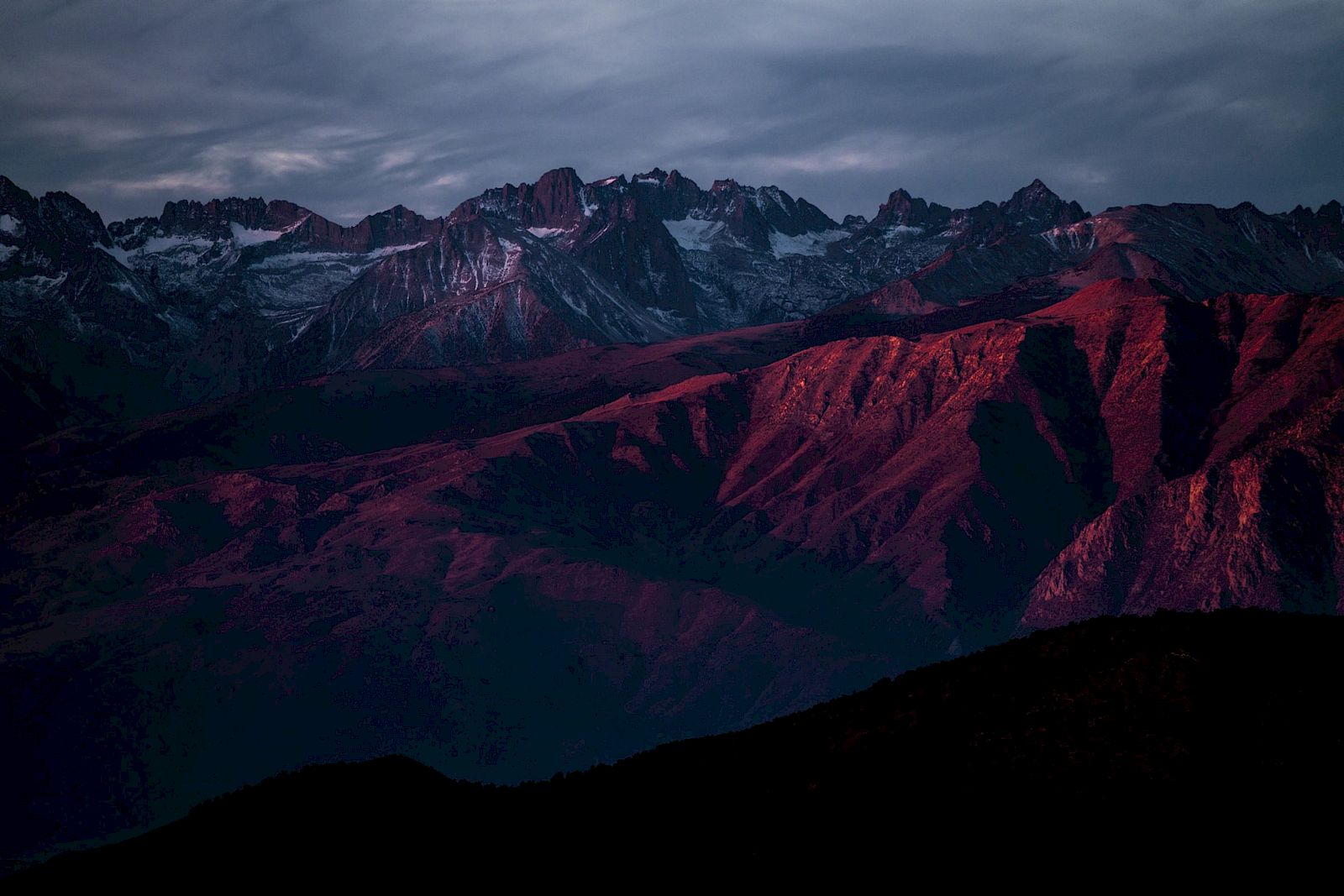 A mountain range under a cloudy sky, with peaks and ridges bathed in a reddish light, possibly during sunset or sunrise.
