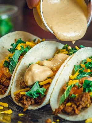 Three tacos filled with ground meat, cheese, lettuce, and tomatoes, being topped with creamy sauce on a plate.