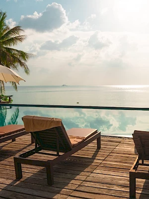 Poolside scene with lounge chairs, umbrellas, and a view of the ocean. Palm trees and a serene atmosphere create a tropical paradise setting.