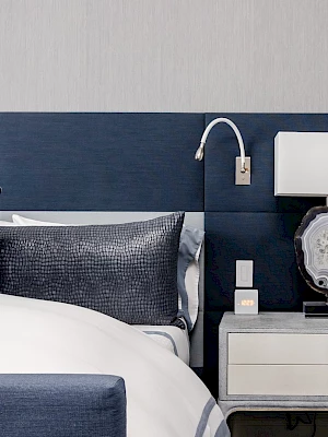 The image shows a modern bedroom with a blue and gray color scheme, featuring a bed, pillows, a nightstand, and a decorative lamp.