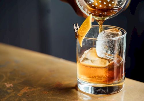 A cocktail is being poured into a glass with ice and a citrus peel garnish, sitting on a countertop.