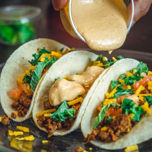 Three tacos with ground meat, lettuce, cheese, and sauce being poured on top, served on a plate.