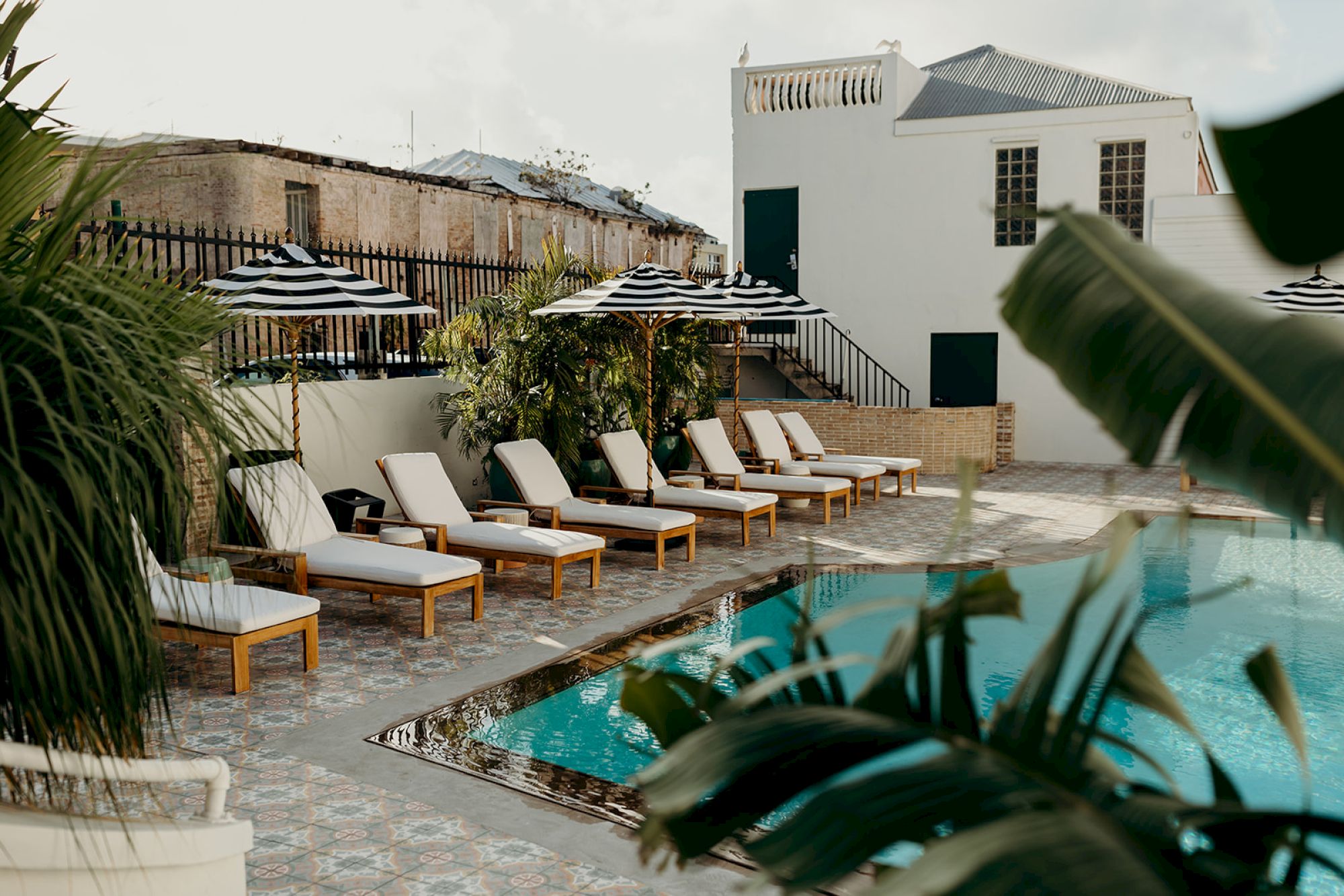A serene pool area with lounge chairs, striped umbrellas, and lush greenery, set beside a building under a clear sky.
