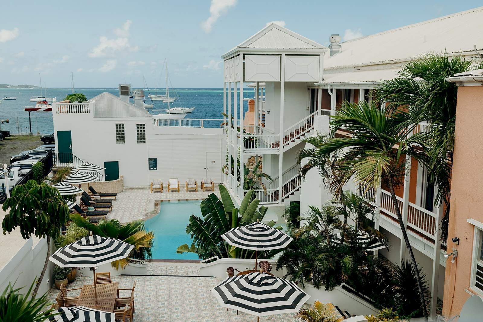 The image shows a coastal hotel with a pool, surrounded by striped umbrellas and palm trees, overlooking the sea with boats anchored nearby.