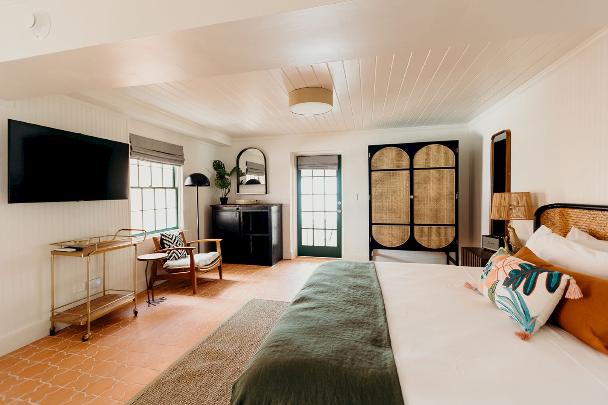 The image shows a cozy bedroom with a bed, TV, armchair, and decorative items, including a mirror and lamp. The decor is modern and stylish.