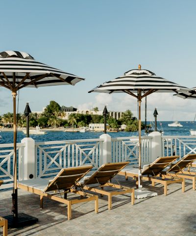 A waterfront view with striped umbrellas and lounge chairs on a deck, overlooking the ocean with boats and a distant shoreline.