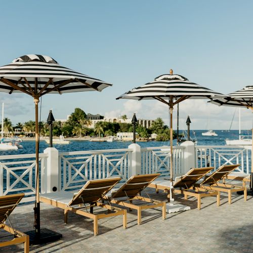 A waterfront view with striped umbrellas and lounge chairs on a deck, overlooking the ocean with boats and a distant shoreline.