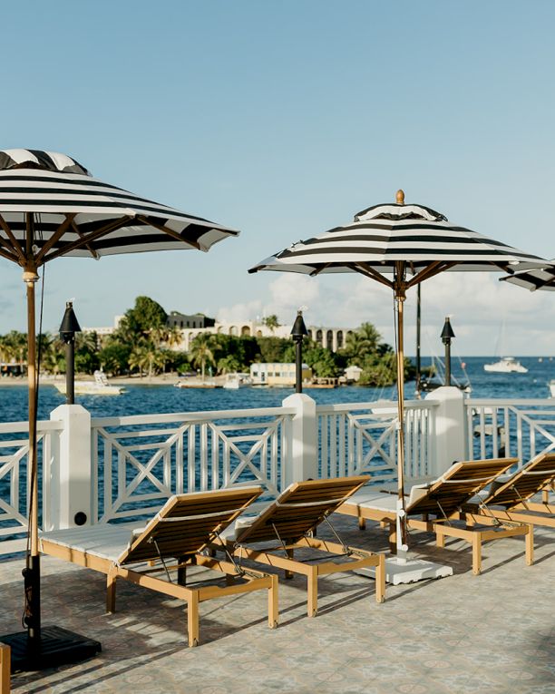A waterfront view with striped umbrellas and lounge chairs on a deck, overlooking the ocean with boats and a distant shoreline.