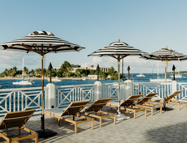 A waterfront view with striped umbrellas and lounge chairs on a deck, overlooking the ocean with boats and a distant shoreline.