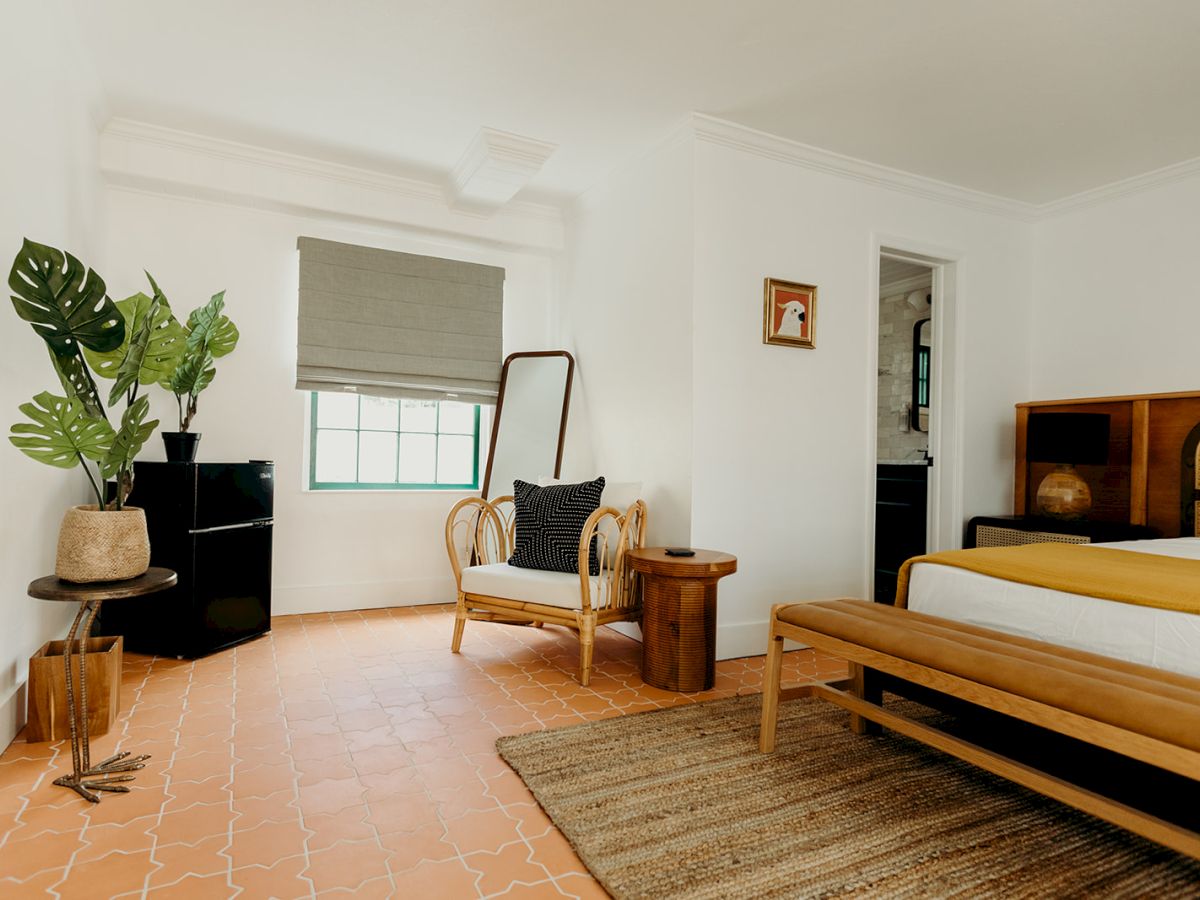 A cozy bedroom with a bed, wicker chair, side table, large plant, and a mirror. It has a terracotta floor and neutral-toned decor.