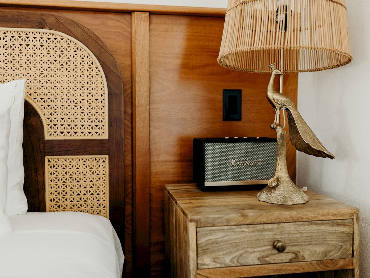 A bedside setup with a wicker headboard, a wooden nightstand, a decorative lamp, and a small speaker placed on the table completes the scene.