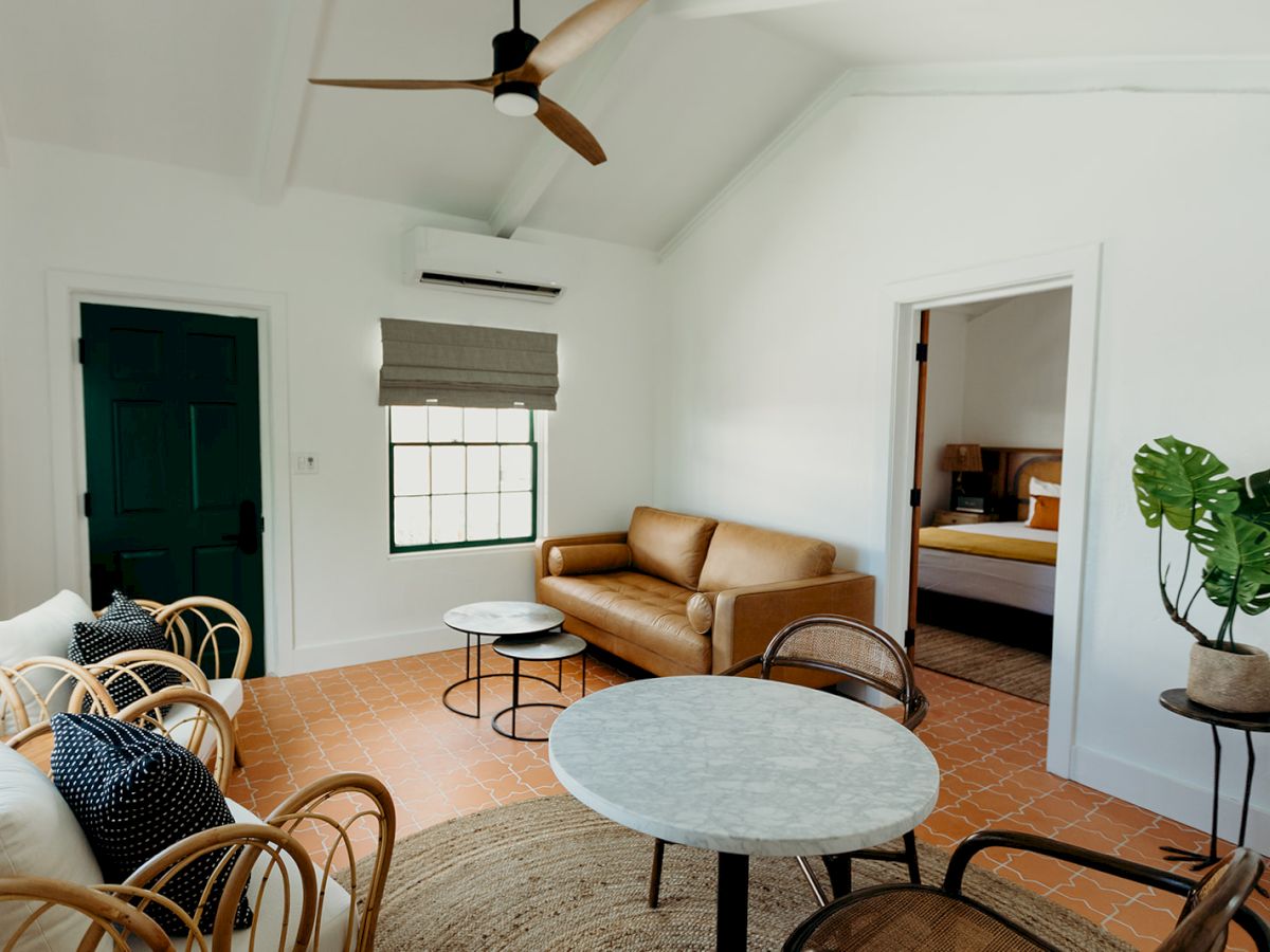 A cozy living room with a sofa, chairs, a ceiling fan, and a table. There's a plant and an open bedroom door to the right.