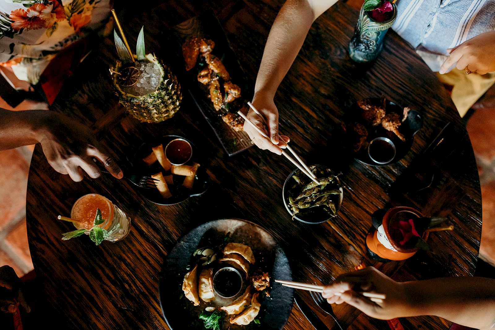 Aerial view of a dining table with diverse food plates and beverages, surrounded by people reaching in with utensils.