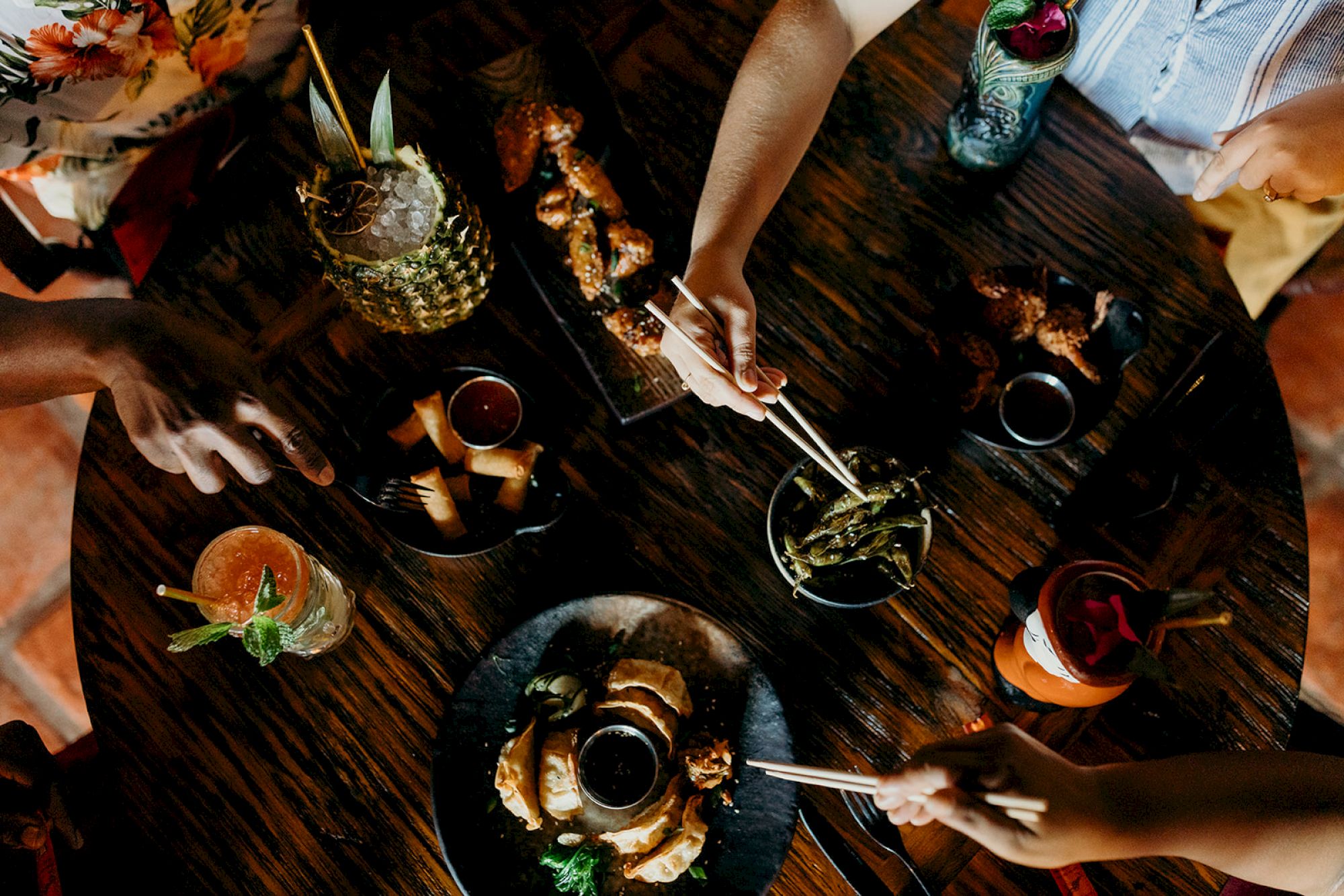 Aerial view of a dining table with diverse food plates and beverages, surrounded by people reaching in with utensils.