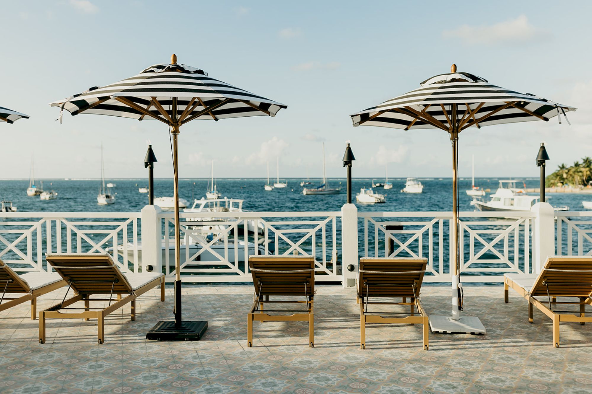 Lounge chairs and striped umbrellas on a deck overlook the sea with sailboats. Calm and scenic view.