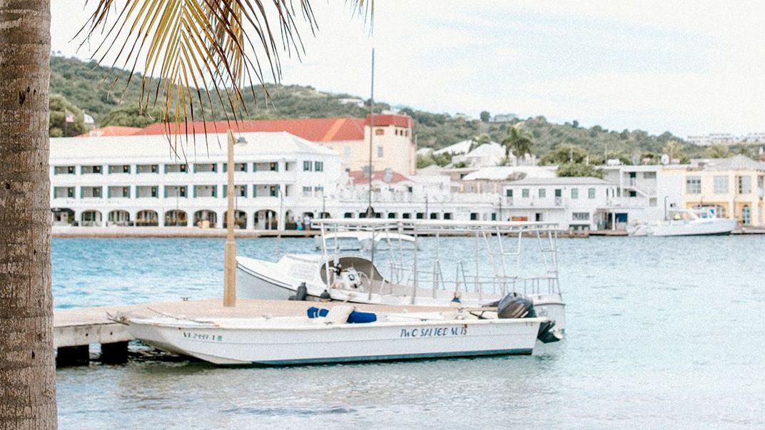 A peaceful waterfront scene with a small boat docked, palm trees, and buildings in the background under a bright sky.
