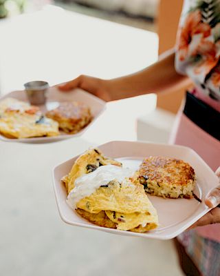 A person holds two plates with omelets and hash browns, wearing a floral shirt and pink apron.