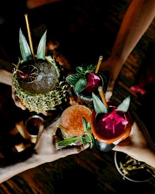 People are holding colorful tropical drinks garnished with fruit and leaves, gathered around a wooden table.