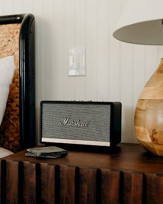 A bed with a wicker headboard is beside a wooden nightstand holding a modern speaker, phone, and lamp, all in a cozy setting.