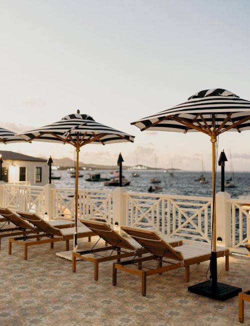 A seaside view with lounge chairs and striped umbrellas lined up by a water's edge, under a partly cloudy sky.