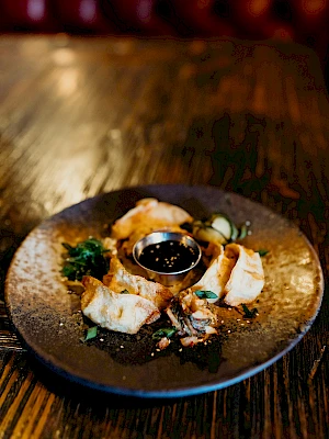 A rustic plate with dumplings surrounds a small bowl of dipping sauce, garnished with herbs, on a wooden table.