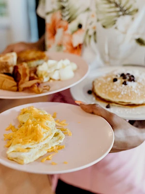 A person is holding three plates with various breakfast foods: pancakes with chocolate chips, scrambled eggs with cheese, and pastries.